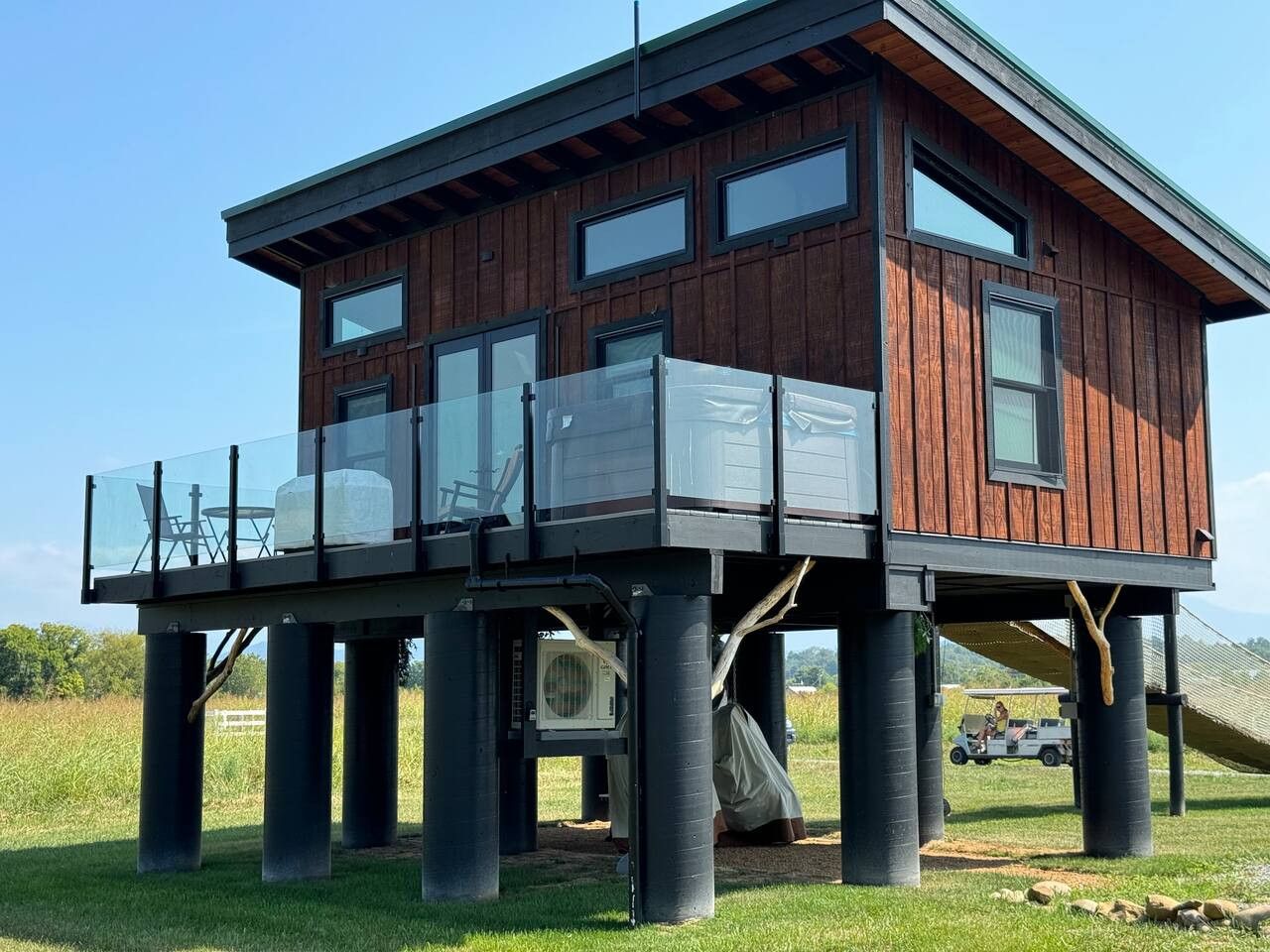 A wooden house with a balcony, supported by pillars, and a vehicle in the background.