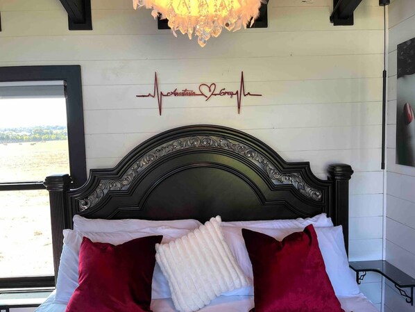 Un lit avec une tête de lit noire, des oreillers blancs et rouges, et une décoration murale en forme de cœur portant le nom « Anastasia Grey ».