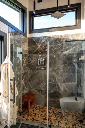 A modern bathroom with a glass shower enclosure, a marble-tiled wall, a wooden stool, and a toilet.