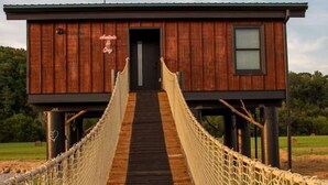 Un pont suspendu en bois mène à un chalet doté d’un toit sombre et d’une petite fenêtre.