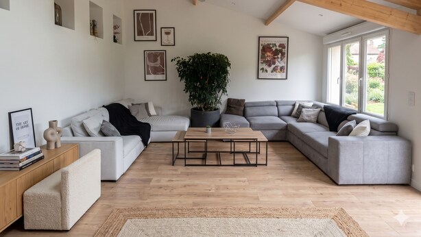 A spacious living room with a high ceiling, wooden beams, a grey sectional sofa, a coffee table, a potted plant, and framed artwork on the walls. - The Crew House Amiens (Cagny)