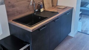 A modern kitchen with a wooden backsplash, black cabinets, a black countertop, a sink, a built-in dishwasher, and a wall-mounted light fixture. - The Crew House Amiens (Cagny)