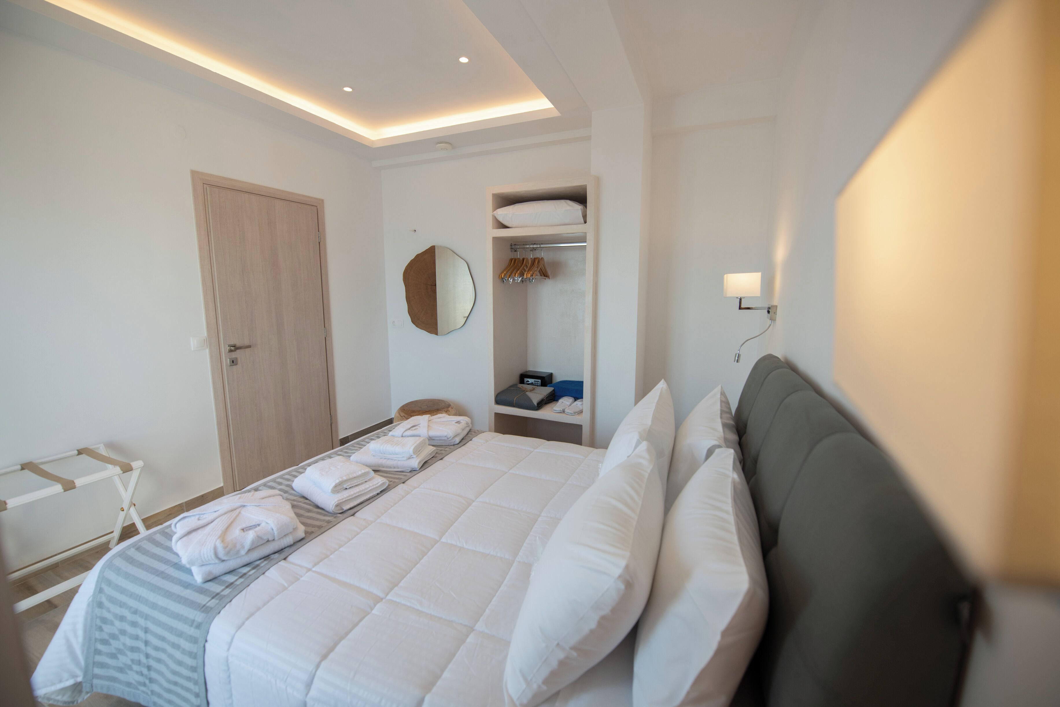 Ein Hotelzimmer mit einem ordentlich bezogenen Bett, einem Kleiderschrank, einem Spiegel und einem Regal mit Handtüchern und einer Uhr.