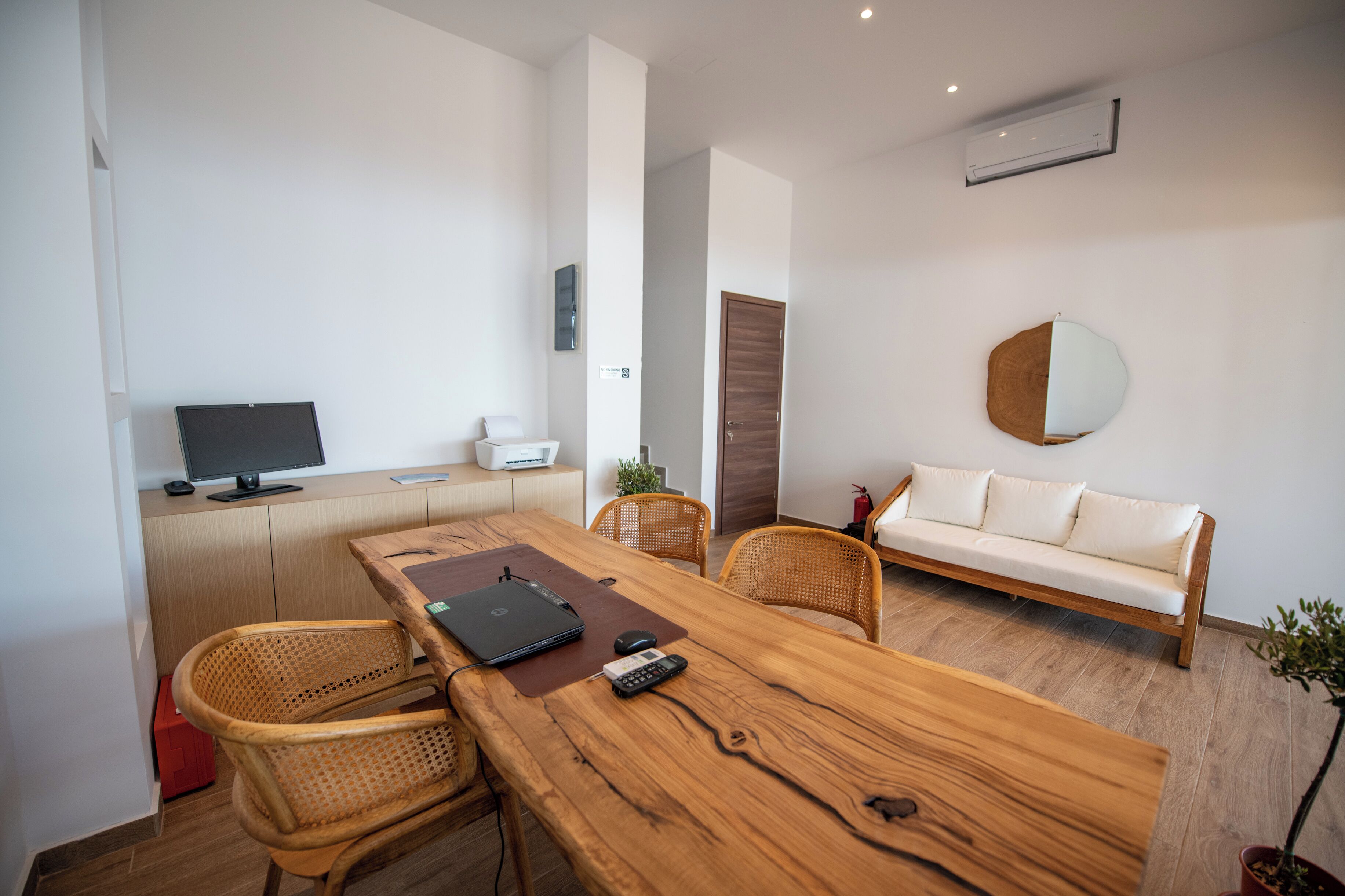 Una habitación moderna con una mesa de comedor de madera, sillas de mimbre, un sofá blanco, un televisor de pantalla plana, una computadora portátil y una planta en maceta.