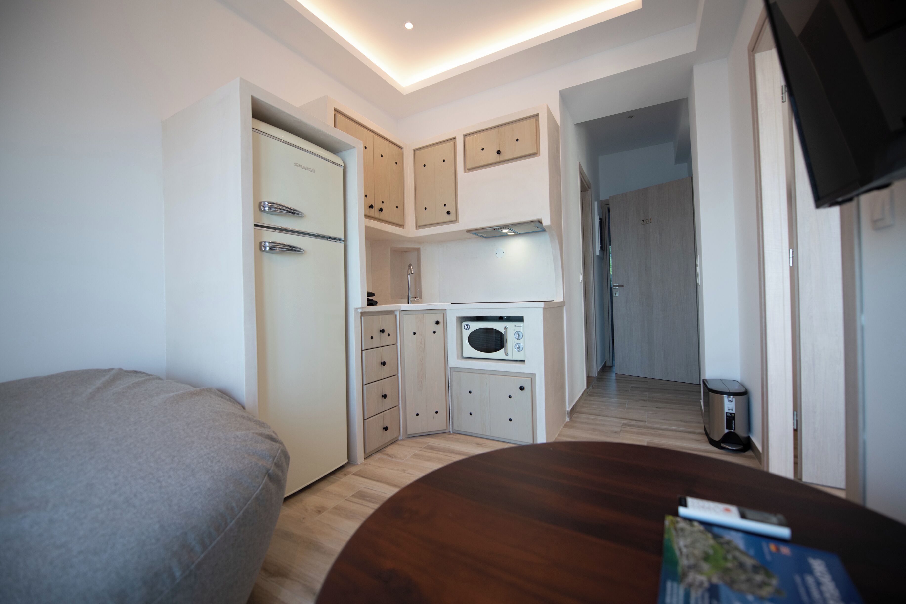 A compact kitchen with a microwave, a refrigerator, and a small cabinet. There is a wooden table with a remote control and a book.