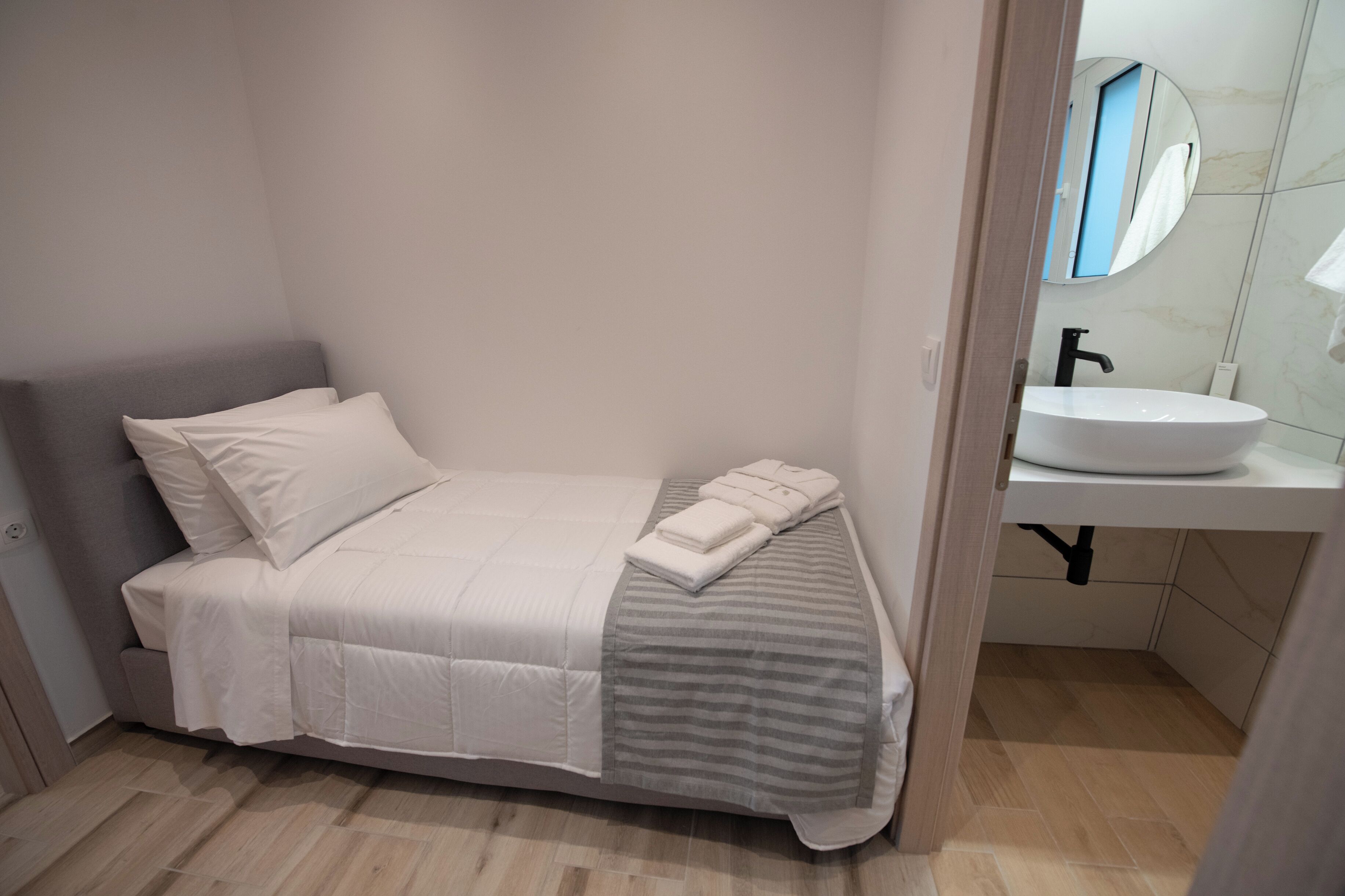 A small, neatly made bed with a white and gray striped blanket, a white pillow, and folded towels on the side. A white sink with a black faucet is visible in the corner, and a mirror above it reflects part of the room.