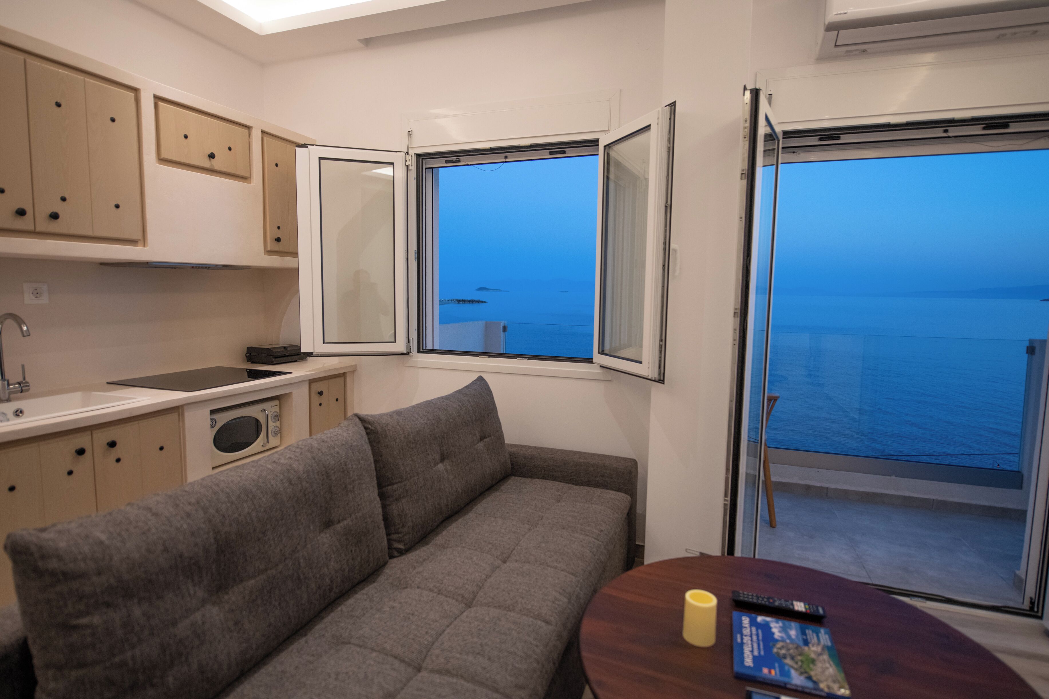 Un espace de vie compact comprenant un coin cuisine, un canapé, une table à manger et une grande fenêtre donnant sur la mer.
