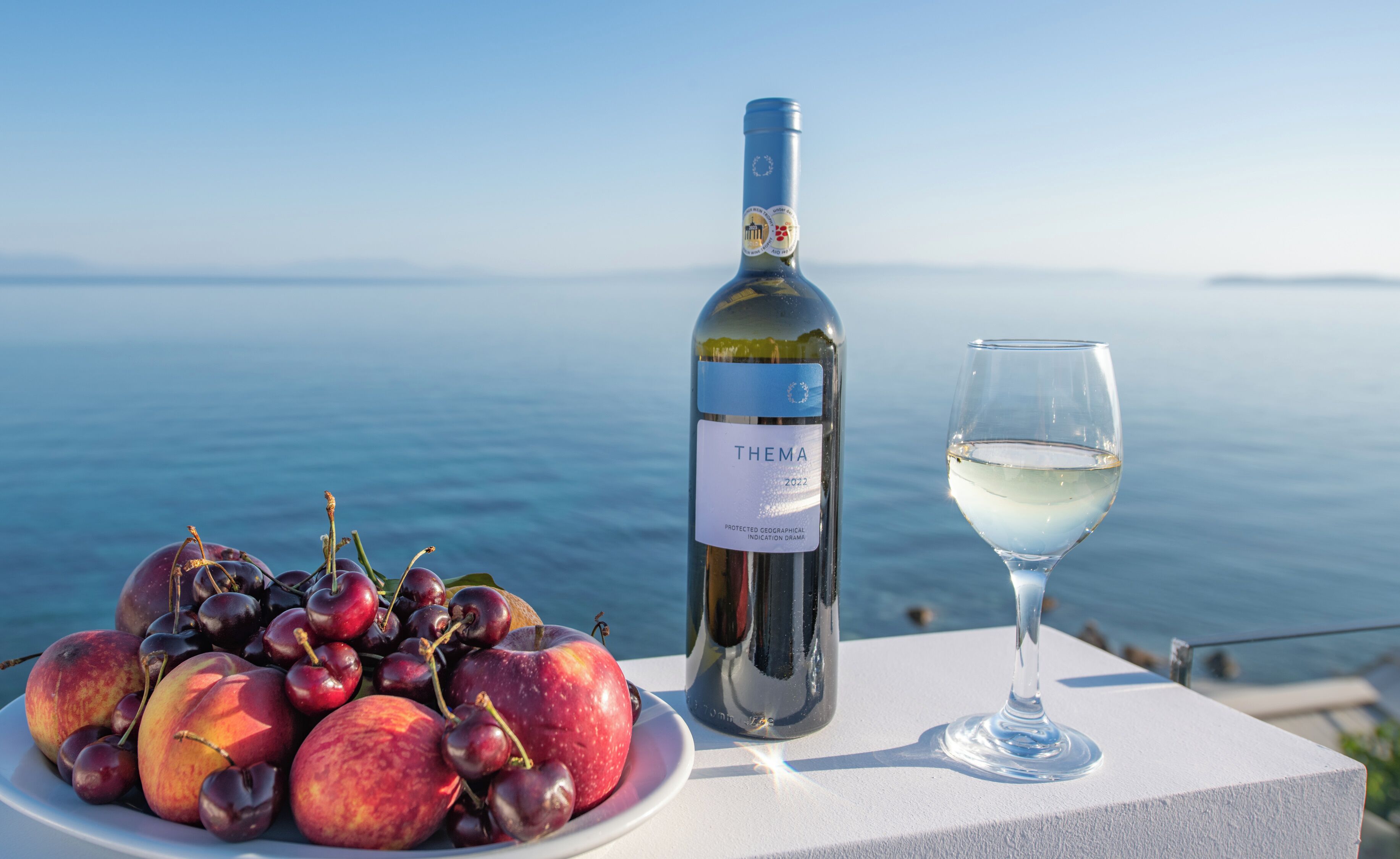 Una ampolla de vi, un got de vi i un plat de fruita sobre una taula amb vista al mar.