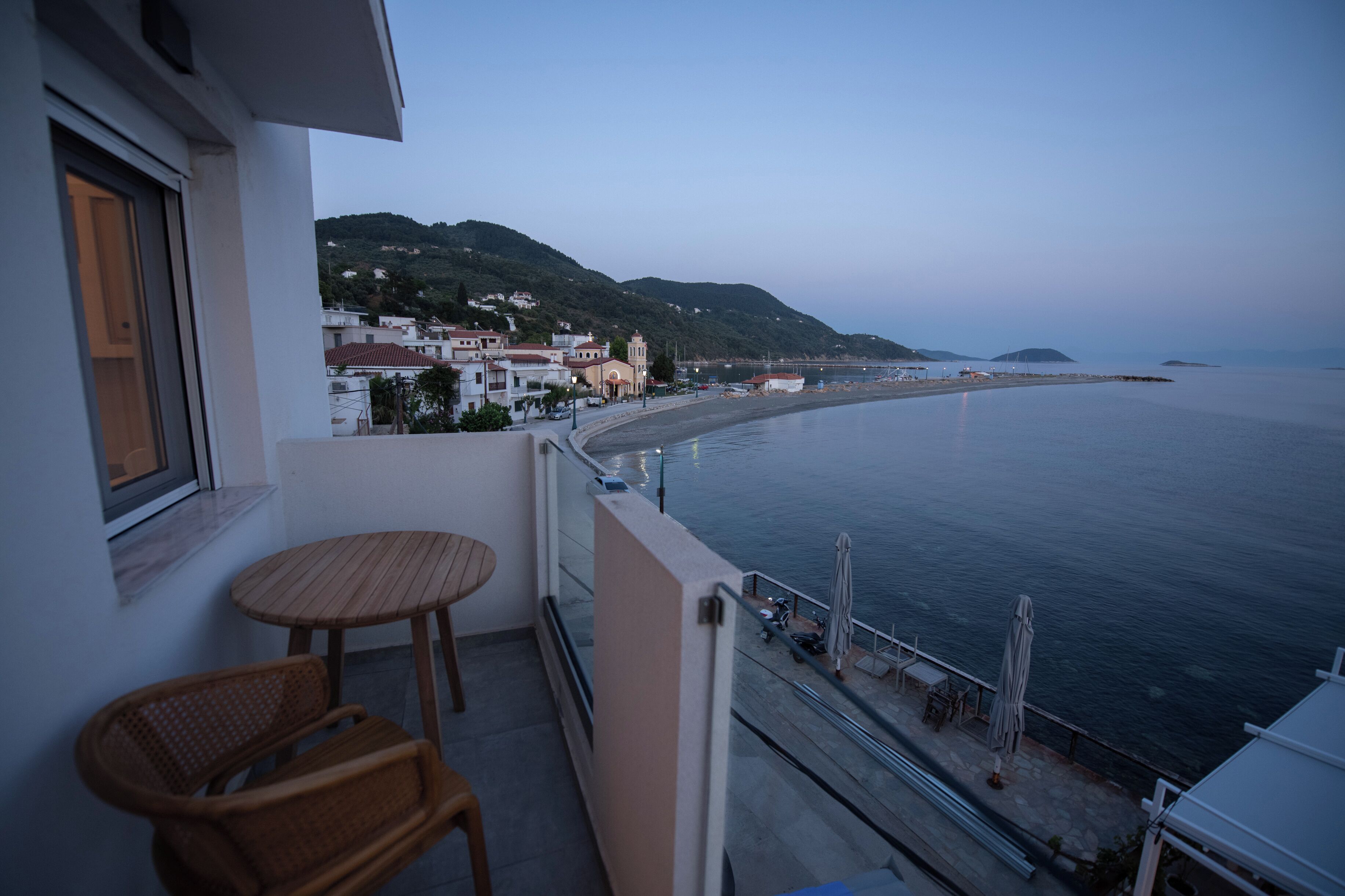 A balcony with a wooden chair and table, overlooking a calm sea and a coastal town with buildings and hills in the background.