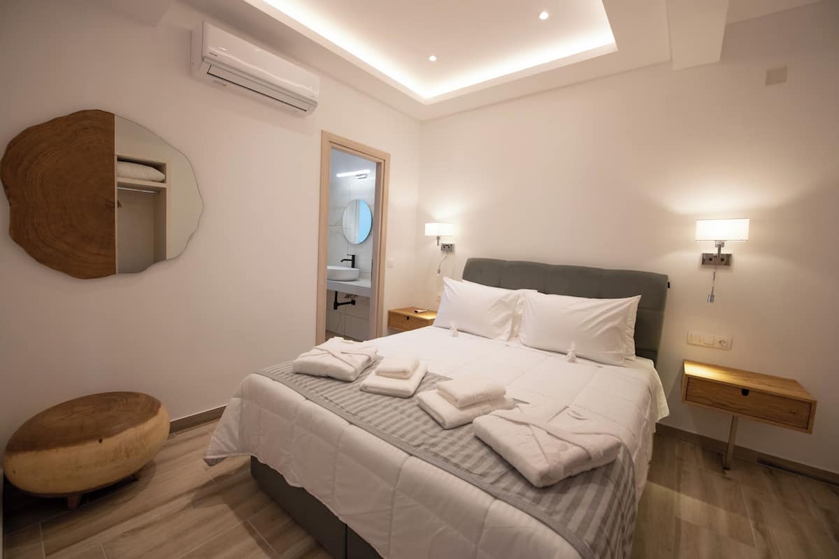 Une chambre d’hôtel moderne avec un grand lit, des tables de chevet, un tabouret rond en bois et un miroir au mur.