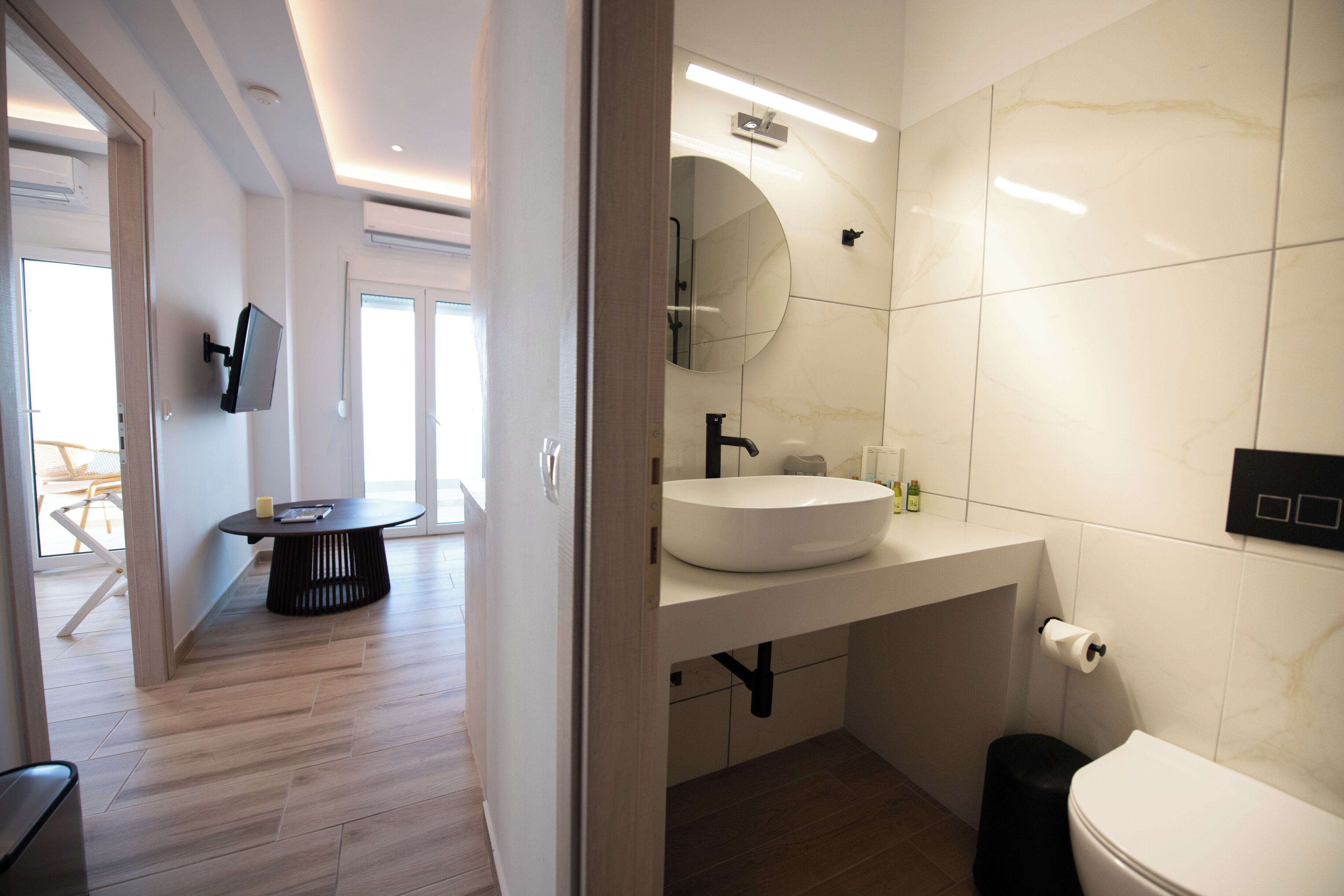Une salle de bain moderne avec un lavabo blanc, un miroir et des toilettes. On trouve une table ronde avec un pied noir dans la pièce adjacente.