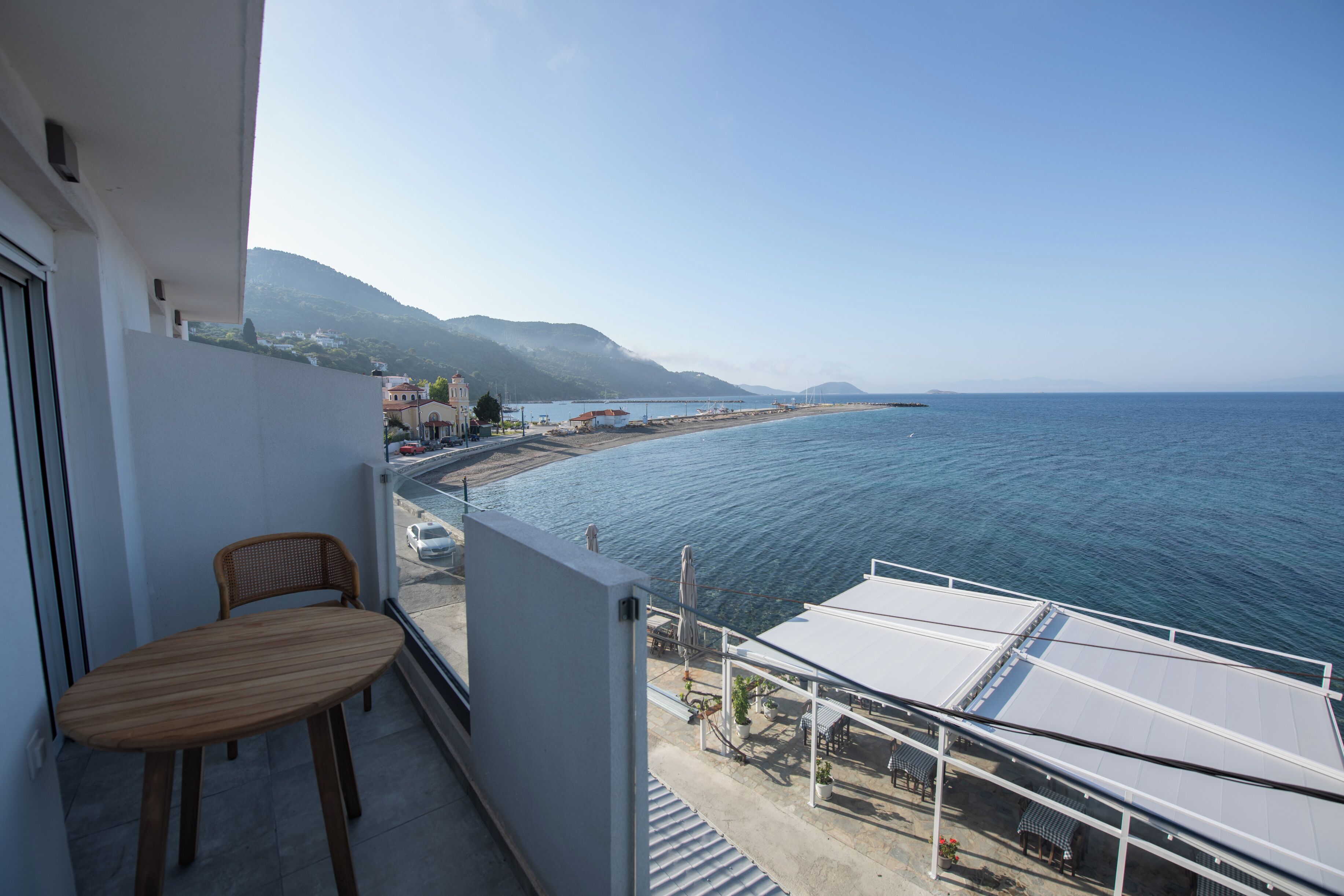A balcony with a wooden table and chairs, overlooking a coastal area with a view of the sea and distant mountains.
