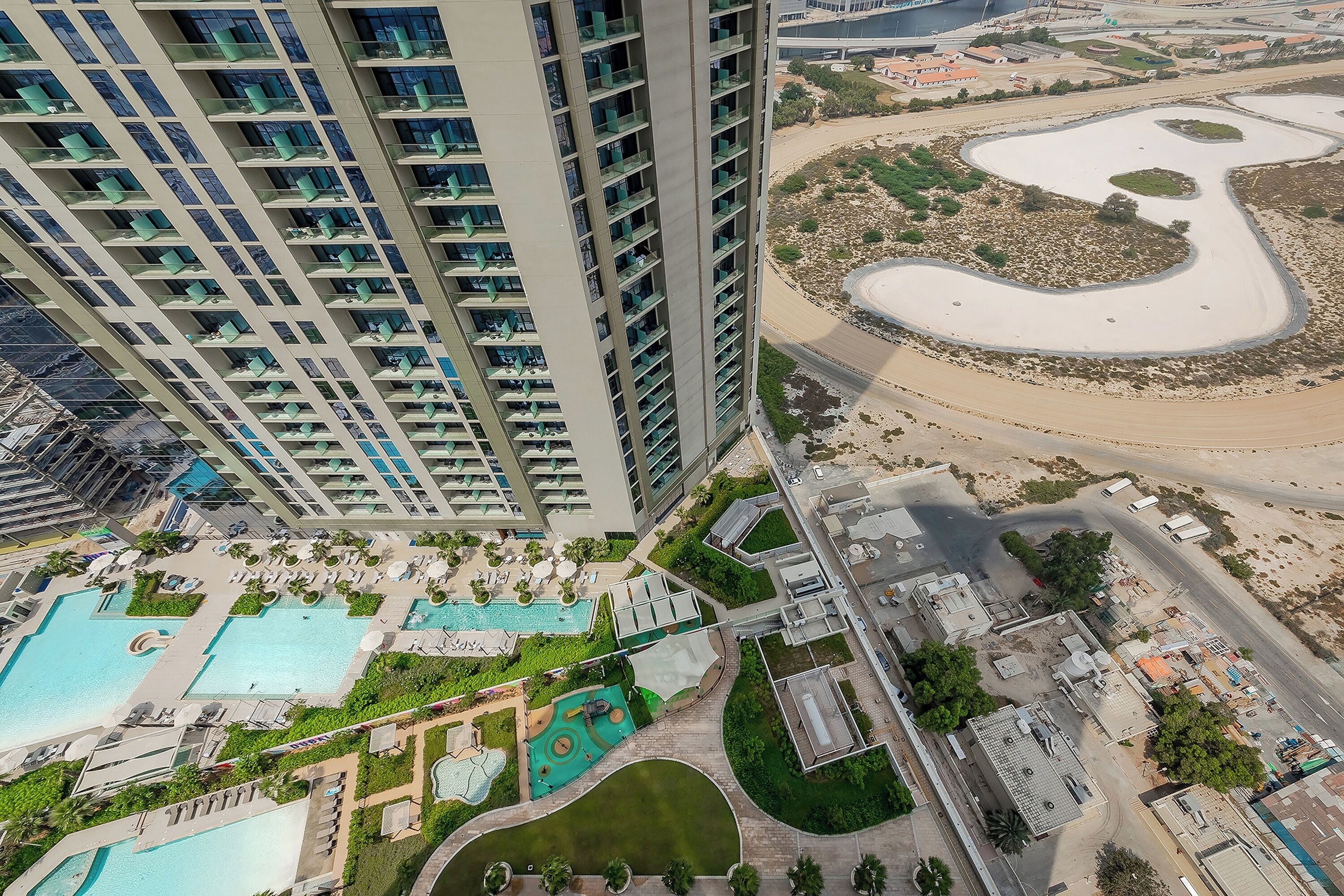A high-rise building with multiple balconies, a landscaped pool area with various water features, and a winding river or canal nearby.