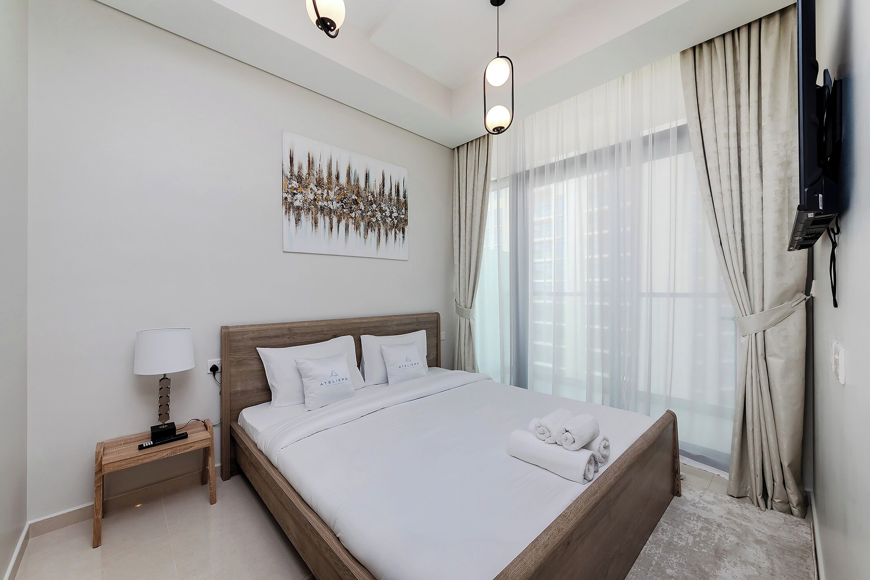 A modern hotel room with a large bed, a bedside table with a lamp, a mounted TV, and a large window with curtains.
