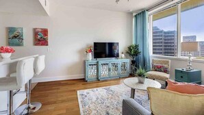 Una sala de estar moderna con televisor de pantalla plana, ventilador de techo, un ventanal con vistas a la ciudad y una zona de comedor con sillas blancas.