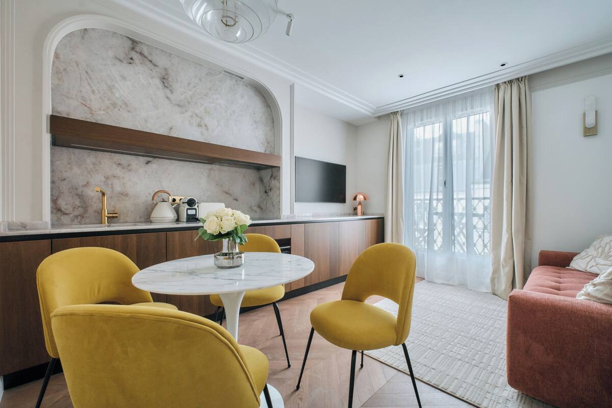 A modern hotel room with a marble accent wall, a kitchen area, a dining table with yellow chairs, and a red sofa.