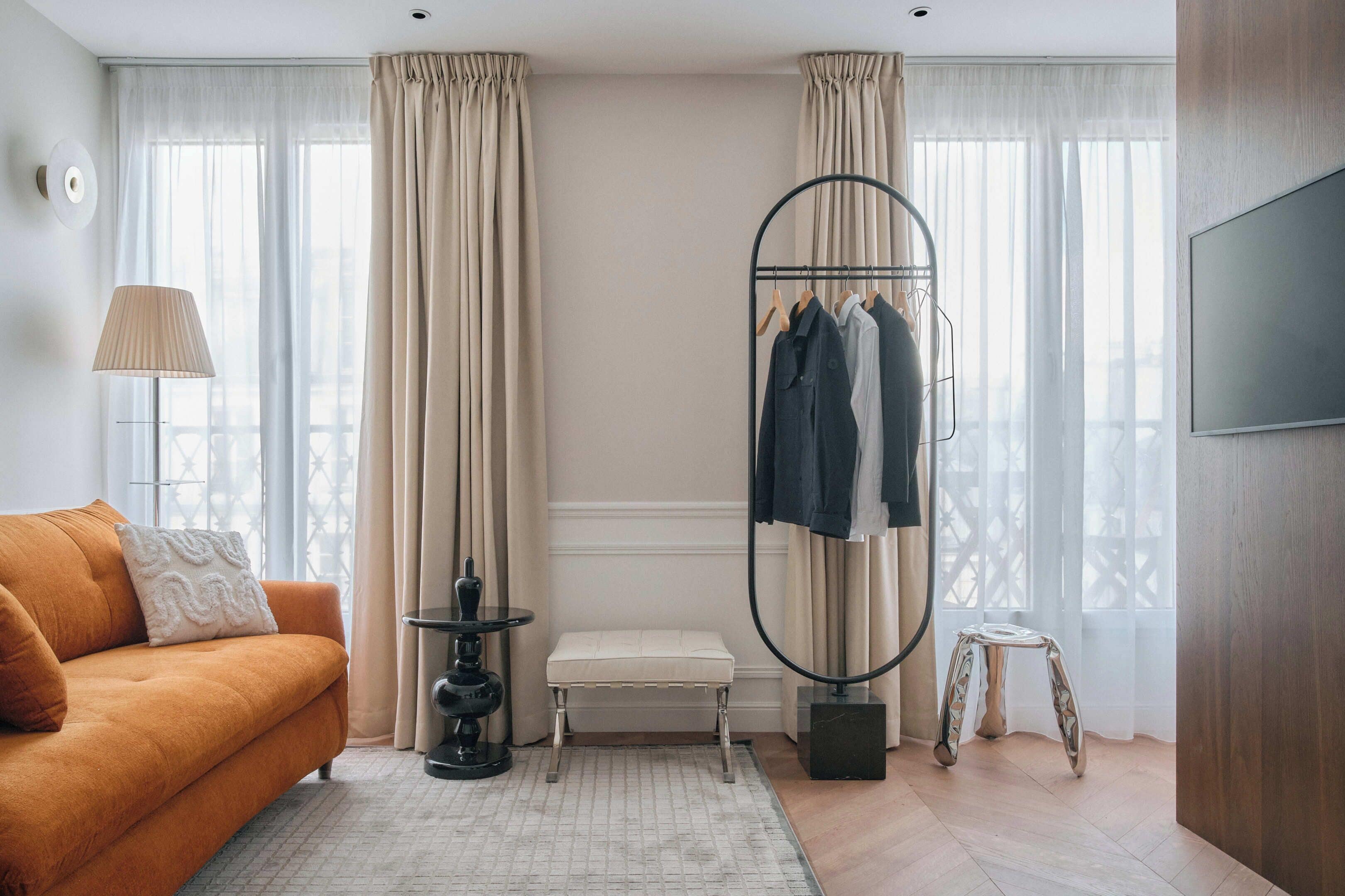 A modern hotel room with a sofa, a hanging clothes rack, a small table, and a TV mounted on the wall.