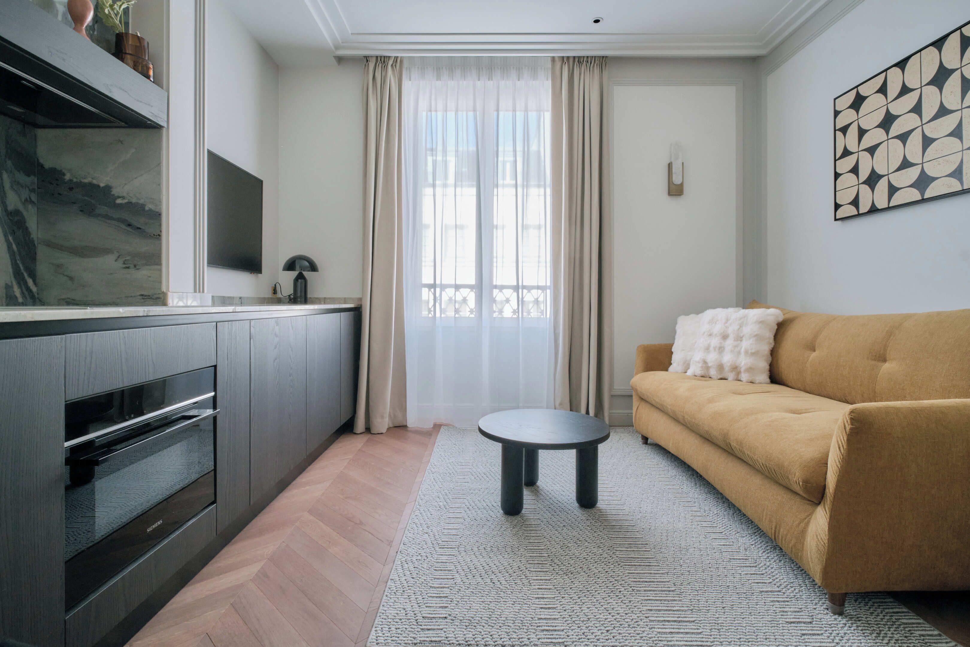 A modern living room with a sofa, a small round table, a kitchen area with a built-in oven, and a large window with sheer curtains.