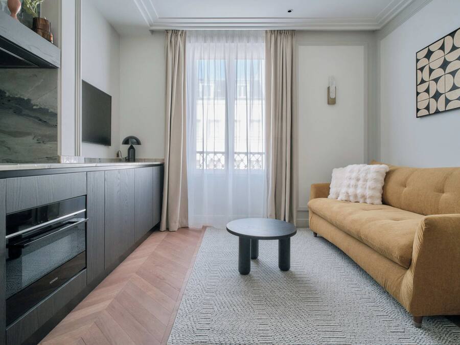 Een moderne woonkamer met een bank, een klein rond tafeltje, een keuken met een inbouwoven en een groot raam met dunne gordijnen.