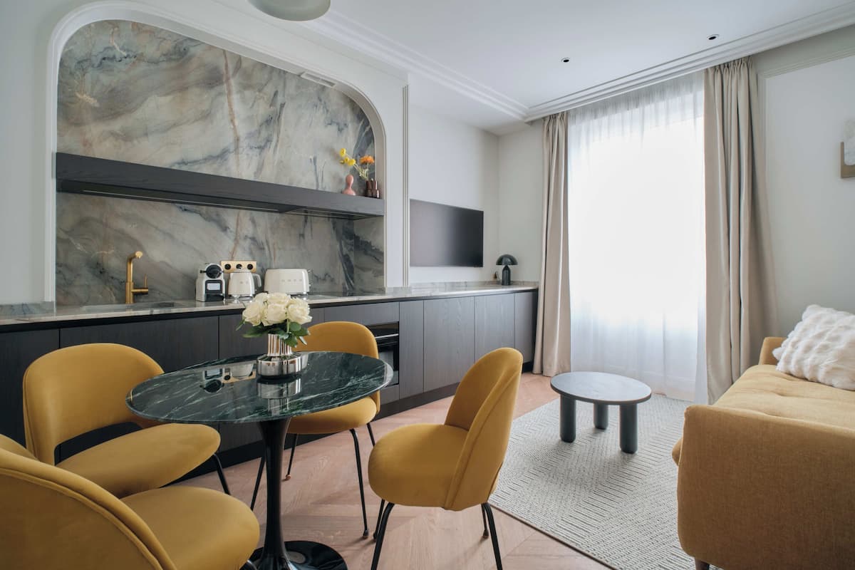 A modern living room with a sofa, a dining table with chairs, a kitchen area with a marble backsplash, and a large window with curtains.
