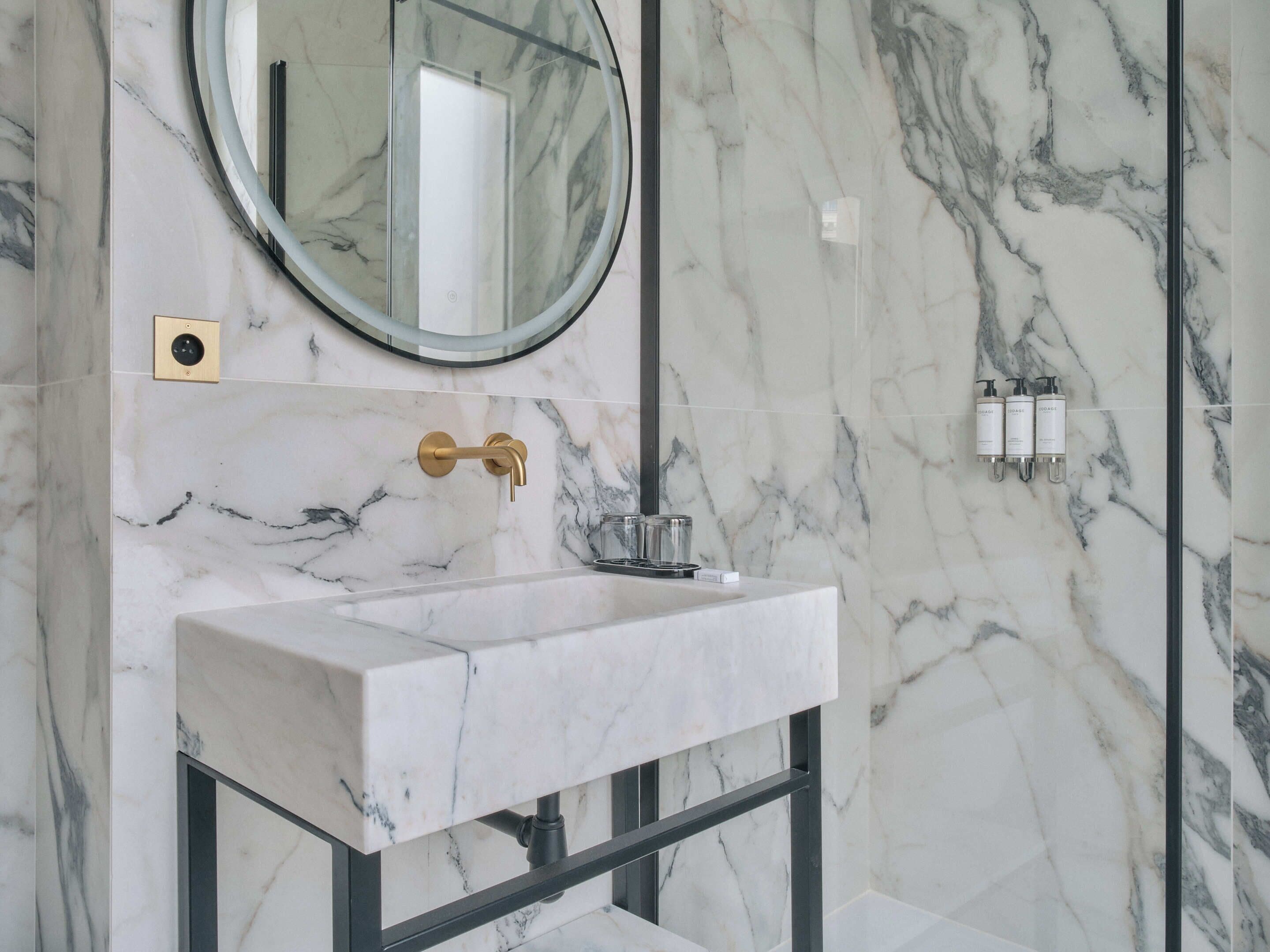 A bathroom with a marble sink, a round mirror, and a gold faucet.