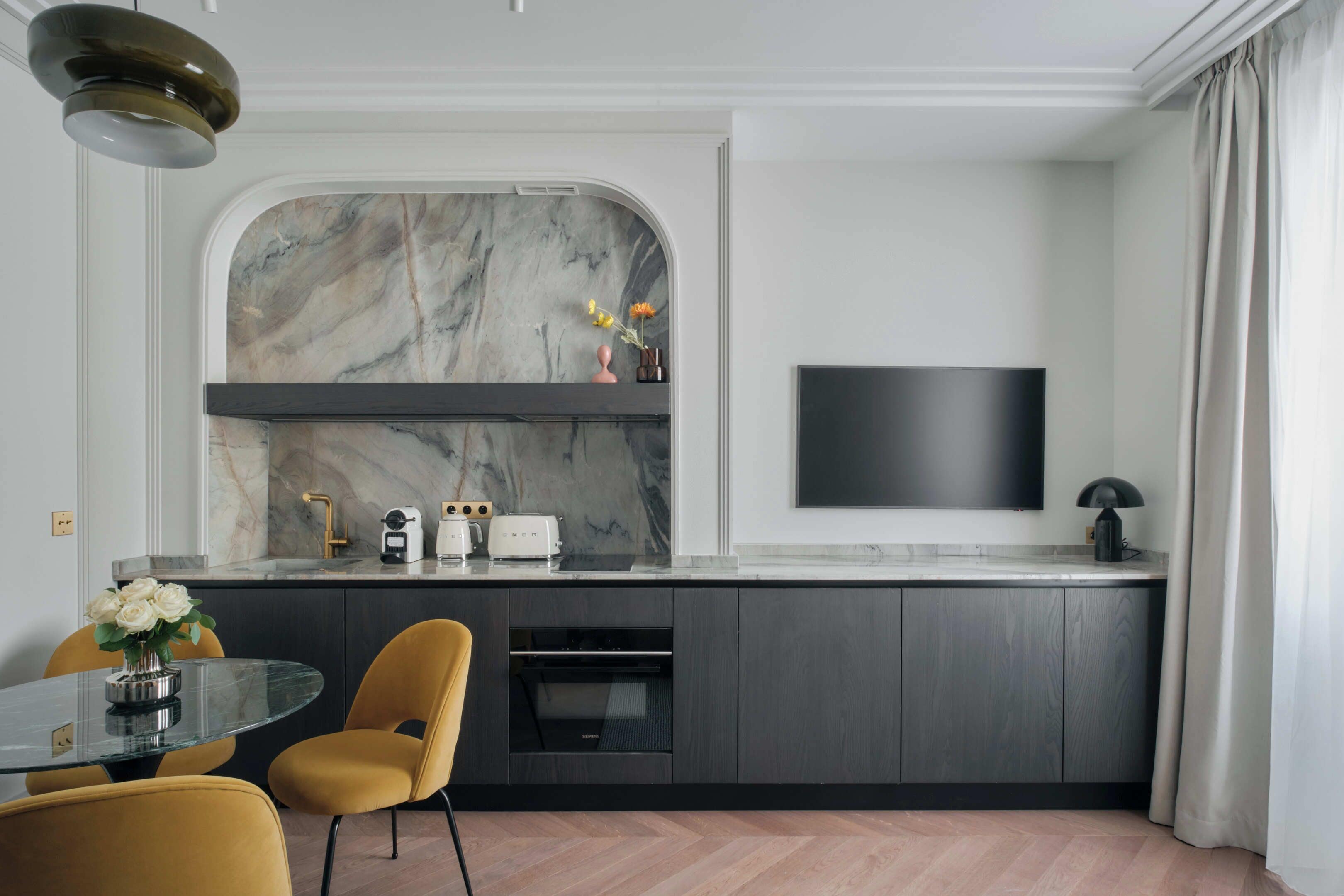A modern kitchen with a marble backsplash, built-in oven, and a flat-screen TV mounted on the wall.