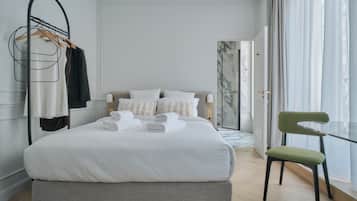 Een net opgemaakt bed met witte lakens en een houten hoofdbord, een groene fauteuil en een glazen tafel bij een groot raam met dunne gordijnen.