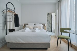 A neatly made bed with white linens and a wooden headboard, a green upholstered chair, and a glass table by a large window with sheer curtains. - Nestay Les Halles (Paris)