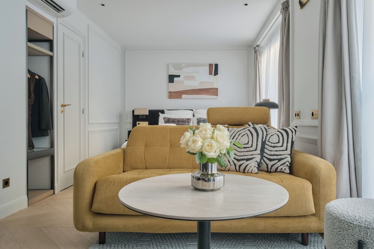 A modern living room with a mustard-colored sofa, a round coffee table with a vase of white flowers, and a minimalist wall art piece.