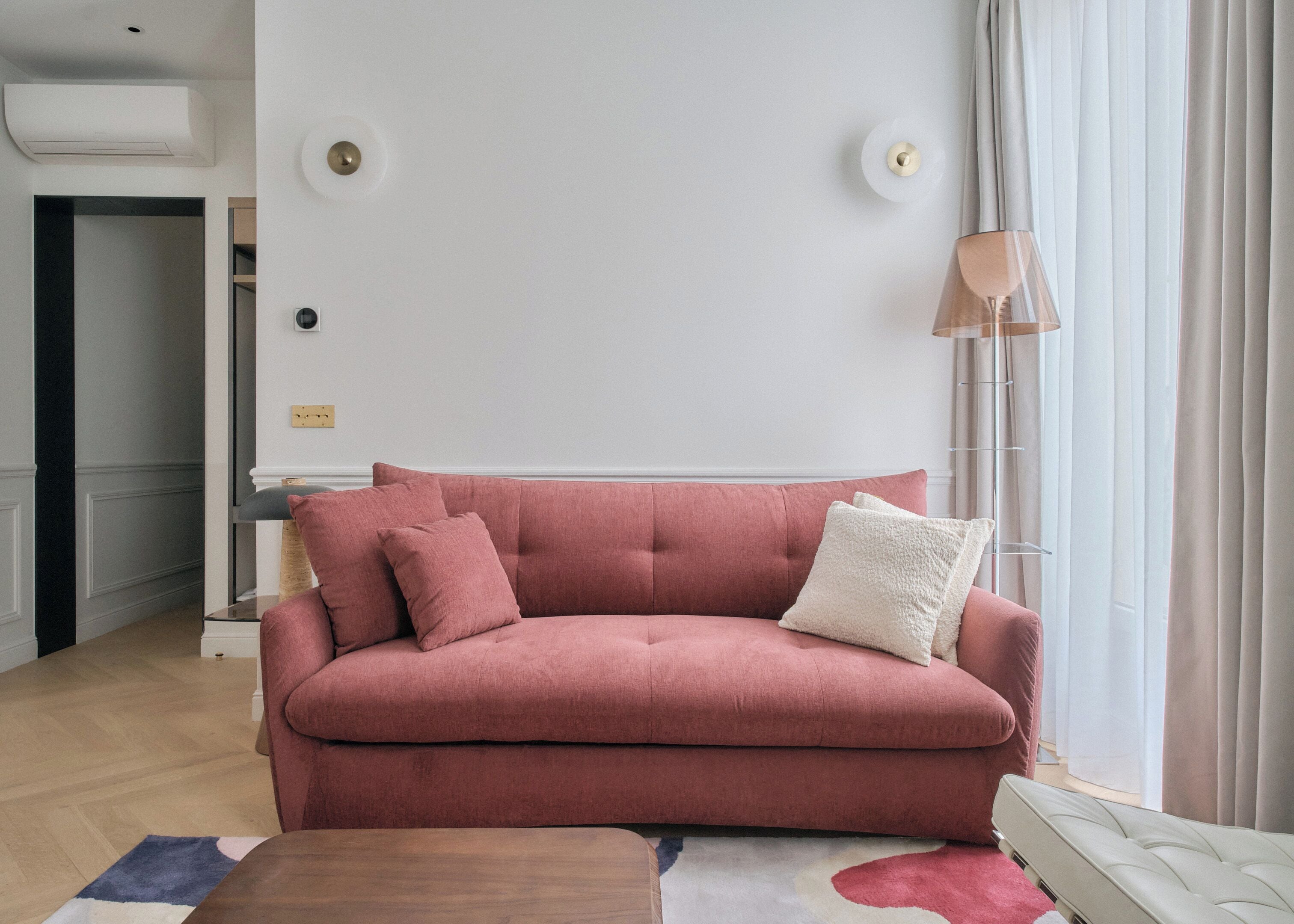 Un salon avec un canapé rose, une petite table et une lampe. Il y a une fenêtre avec des rideaux et une porte qui donne sur une autre pièce.