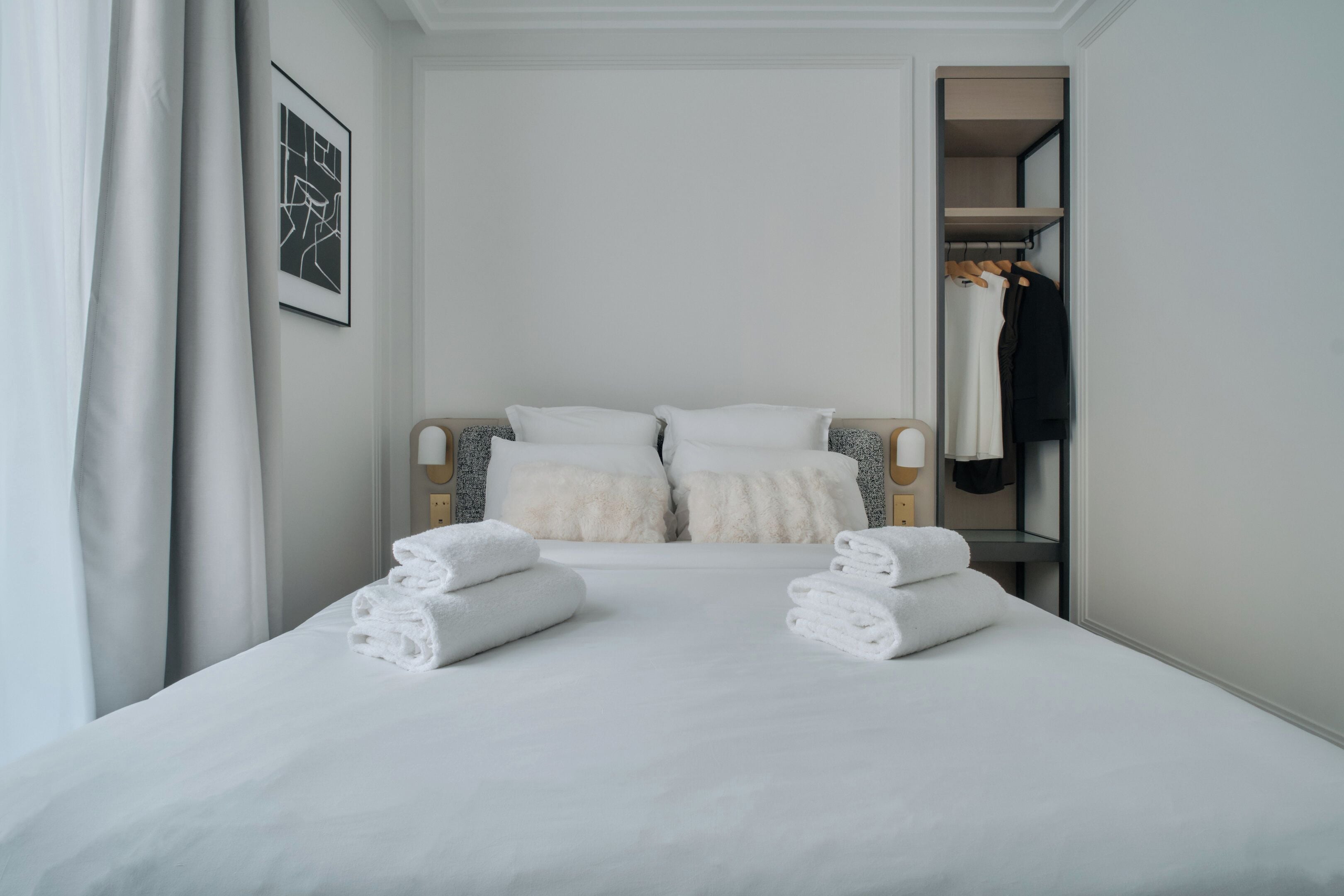 A neatly made bed with white linens and pillows, a wooden headboard, and a white wall with a framed picture.