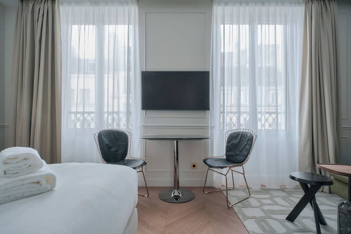 A hotel room with a bed, two chairs, a small table, a TV, and large windows with curtains.