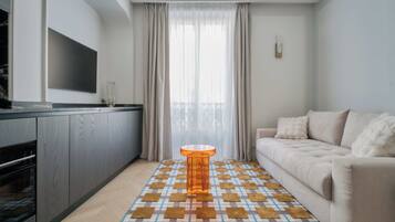 Ein modernes Wohnzimmer mit einer Couch, einem gemusterten Teppich, einem kleinen orangefarbenen Tisch und einer Küchenzeile inklusive Fernseher und Schränken.