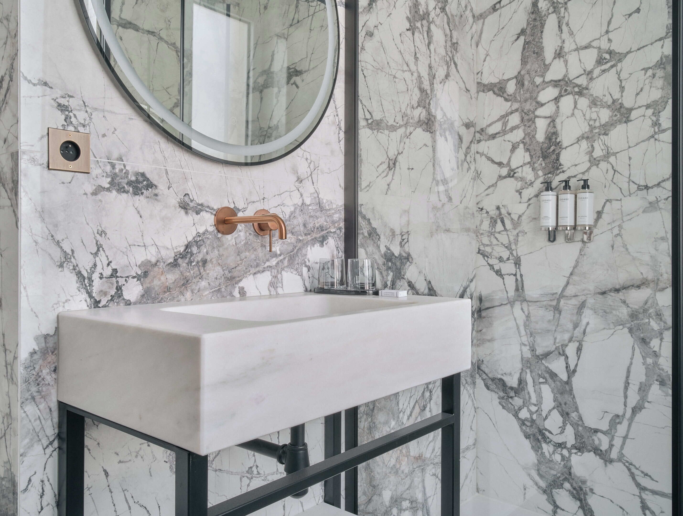 A bathroom with a marble backsplash, a round mirror, a white sink, and black metal frames.