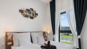 A neatly made bed with white linens and brown towels, a wall-mounted light fixture, and a view of greenery through a window with dark curtains. - Javorná Lodge by Interhome (Javorná)