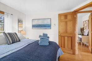 A bedroom with a bed, a wooden door, a desk, and a painting of the ocean. - Sand Cove Cottage-New! (Gouldsboro)