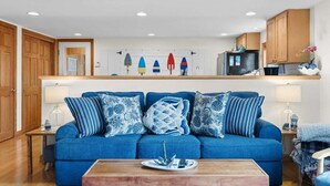 A living room with a blue sofa, patterned cushions, a wooden coffee table, and a kitchen area with wooden cabinets and a refrigerator. - Sand Cove Cottage-New! (Gouldsboro)