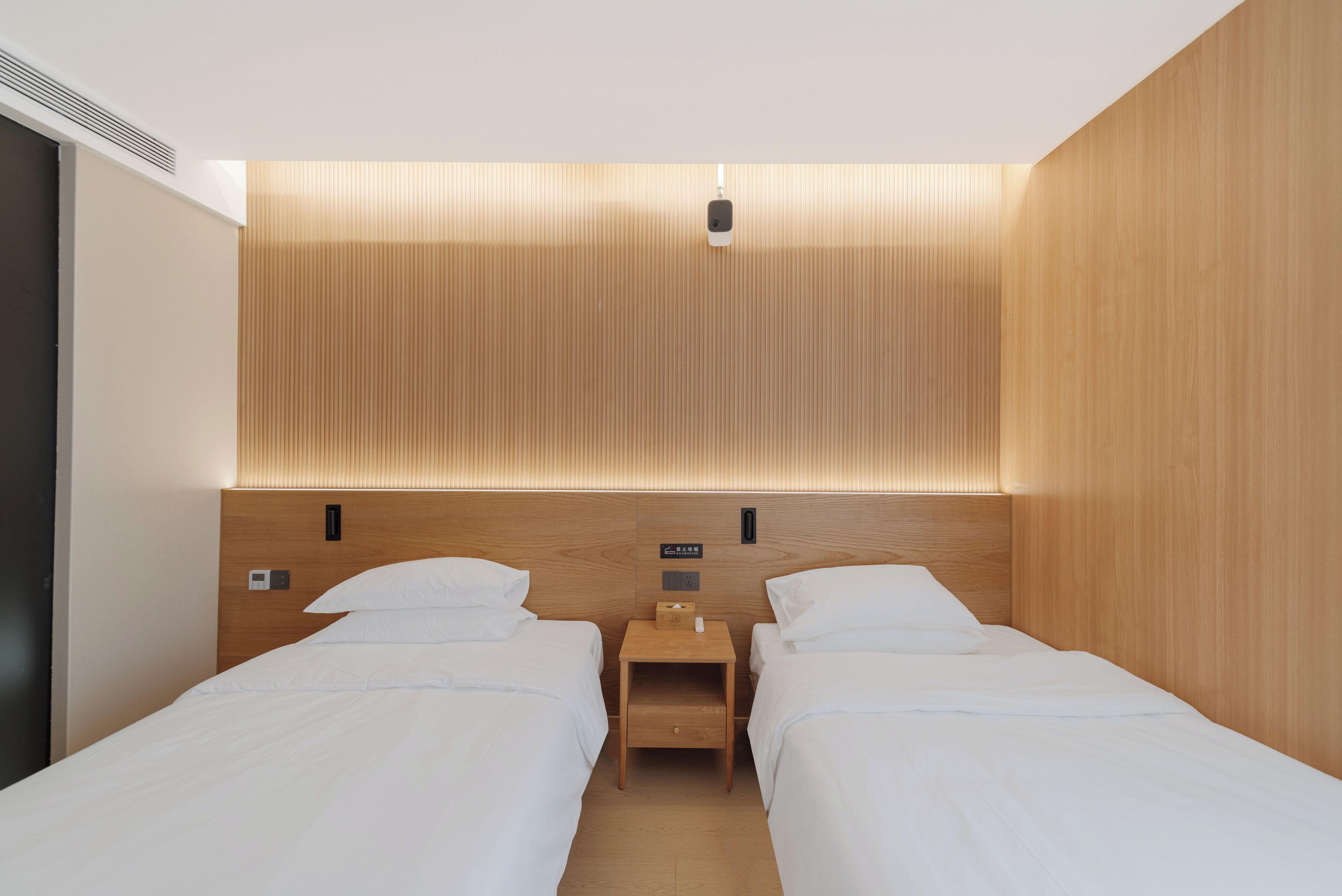 A hotel room with two single beds, a wooden headboard, a bedside table with a lamp, and a small shelf.