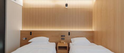 A hotel room with two single beds, a wooden headboard, a bedside table with a lamp, and a small shelf.