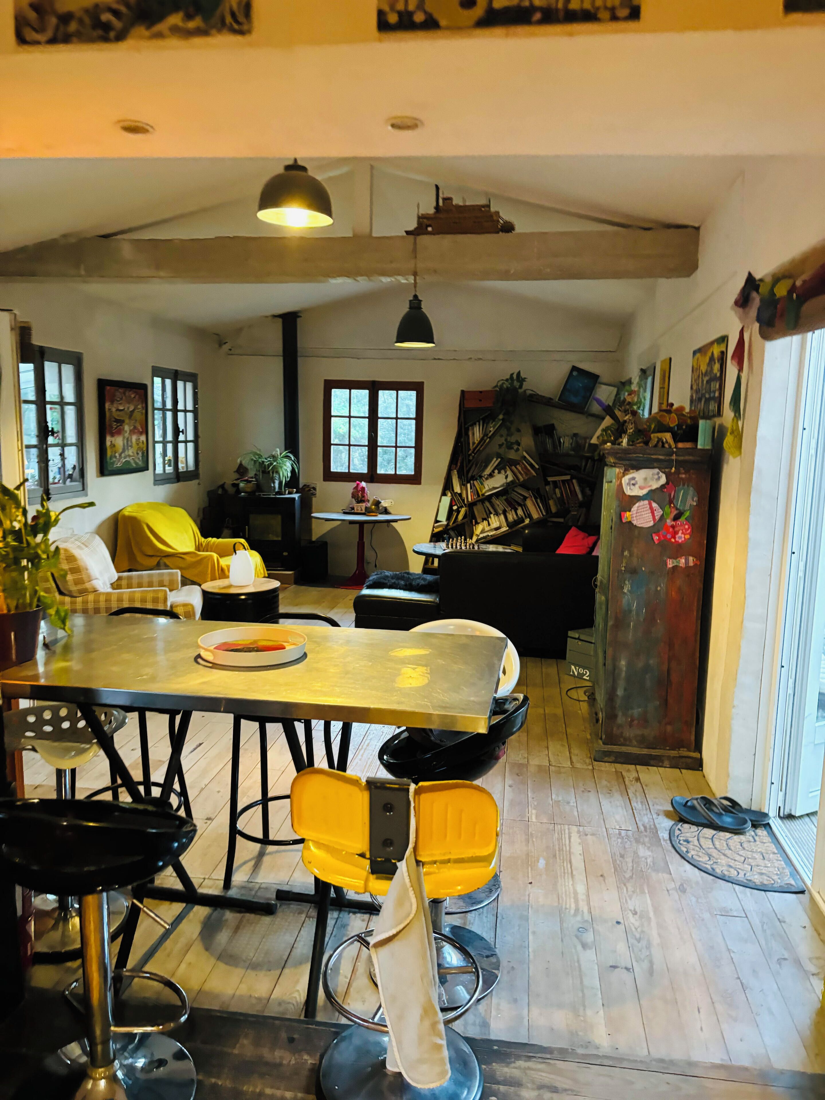 Ein Essbereich mit Holztisch, gelben Stühlen und einem schwarzen Barhocker. Es gibt ein Bücherregal mit Büchern und Dekoration, eine Topfpflanze und ein Fenster mit Vorhängen.