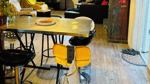 Ein Essbereich mit Holztisch, gelben Stühlen und einem schwarzen Barhocker. Es gibt ein Bücherregal mit Büchern und Dekoration, eine Topfpflanze und ein Fenster mit Vorhängen.