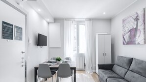 A modern living room with a grey sofa, a dining table with chairs, a TV mounted on the wall, and a large window with sheer curtains. - Easylife -Modern and elegant in the heart of Milan (Milano)
