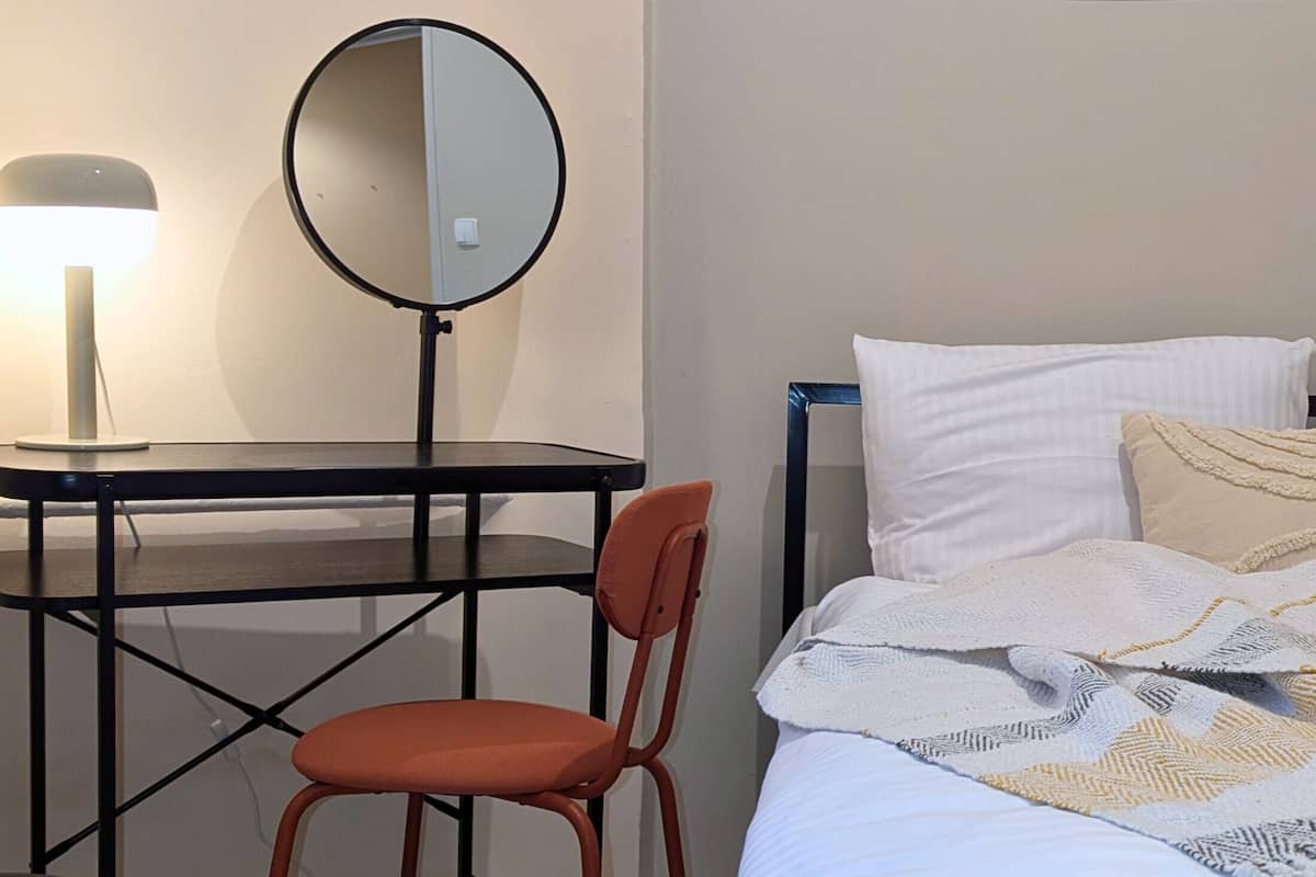 A room with a bed, a desk with a round mirror, a chair, and a lamp.