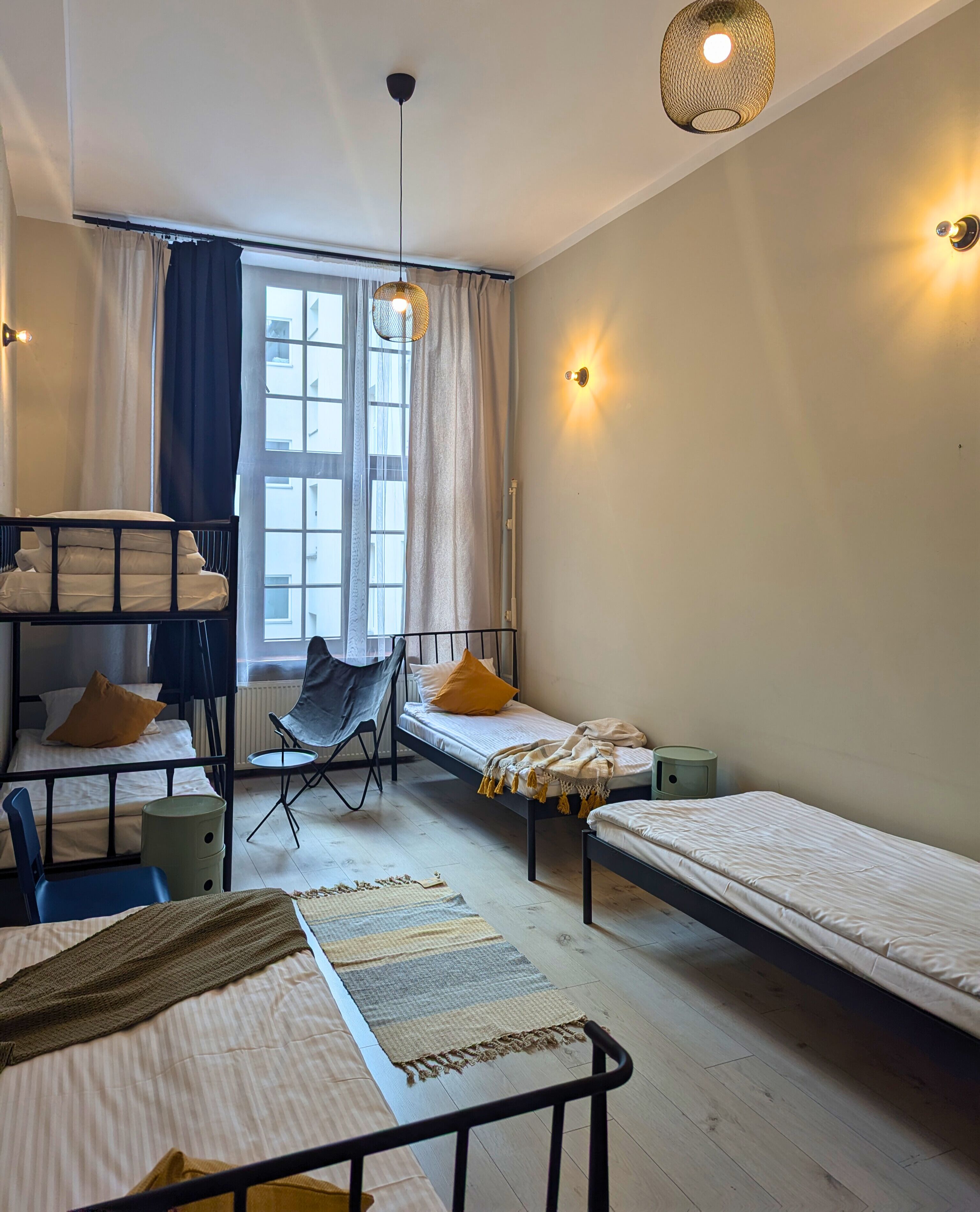 Quarto de dormitório com beliches, uma cadeira dobrável, uma pequena mesa e uma janela com cortinas.