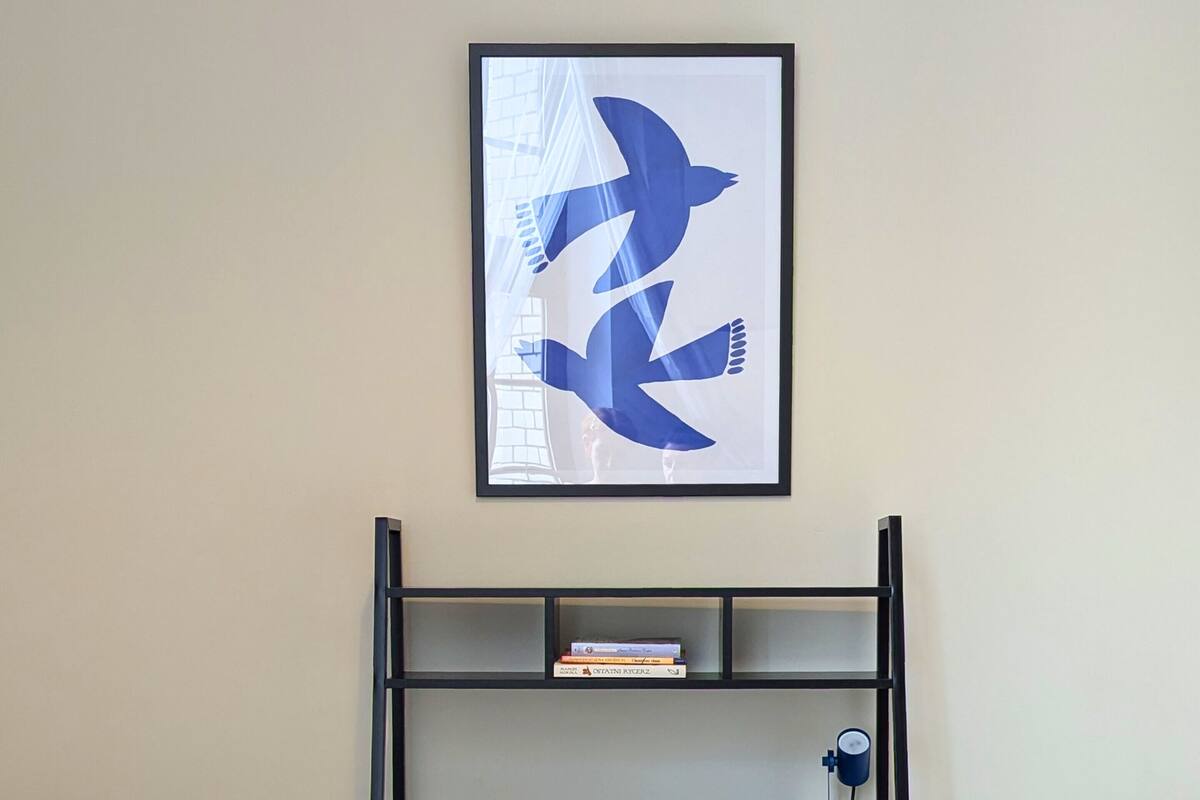 A dormitory room with a bunk bed, a desk with a chair, a framed abstract artwork, and a bookshelf.