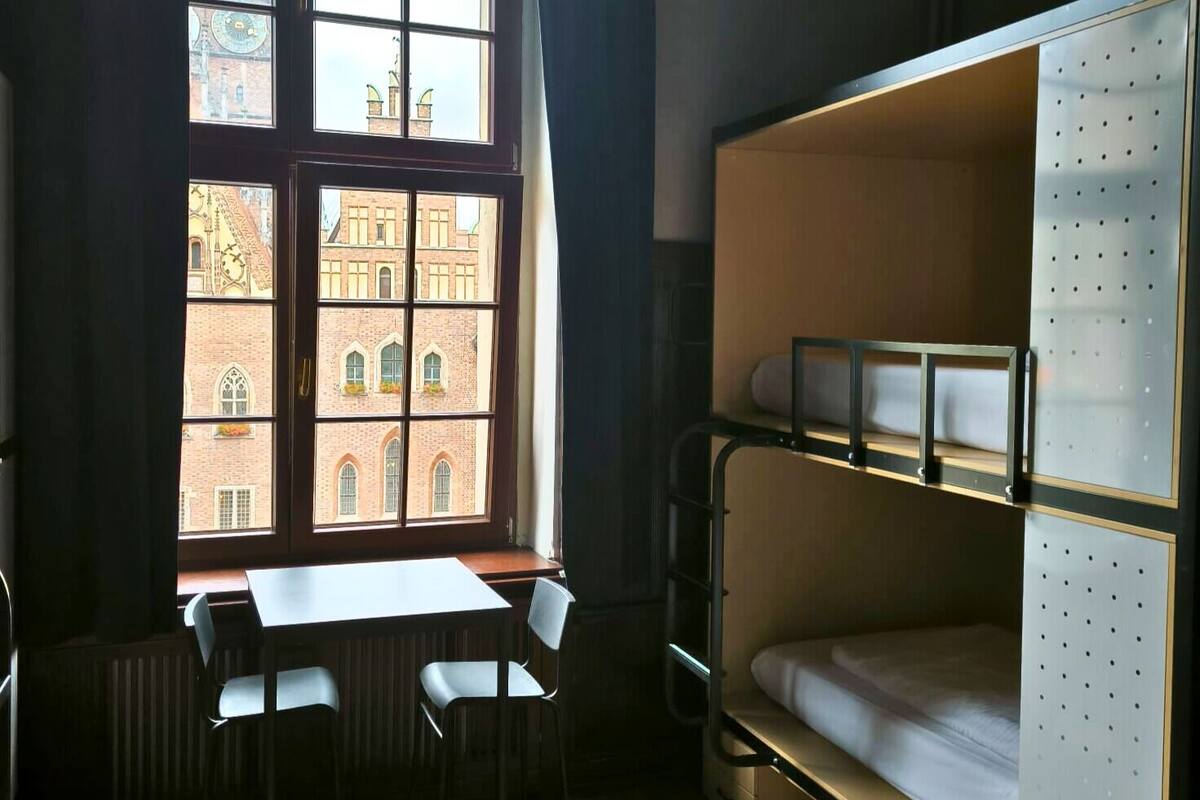 A dormitory room with bunk beds, a table, two chairs, and a window with a view of a historic building.