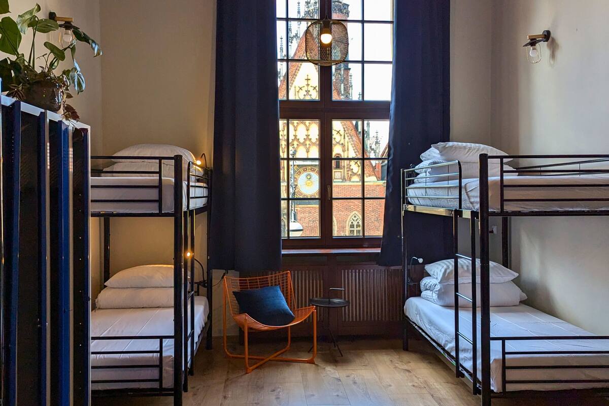 A dormitory room with bunk beds, a wicker chair, and a window with a view of a building.