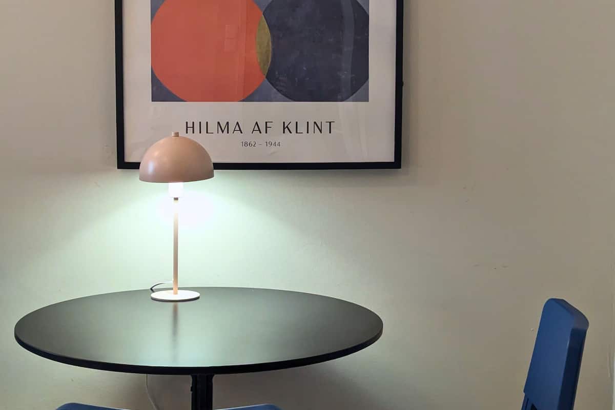 A framed abstract artwork by Hilma af Klint on a wall, a round table with a lamp, and two blue chairs.