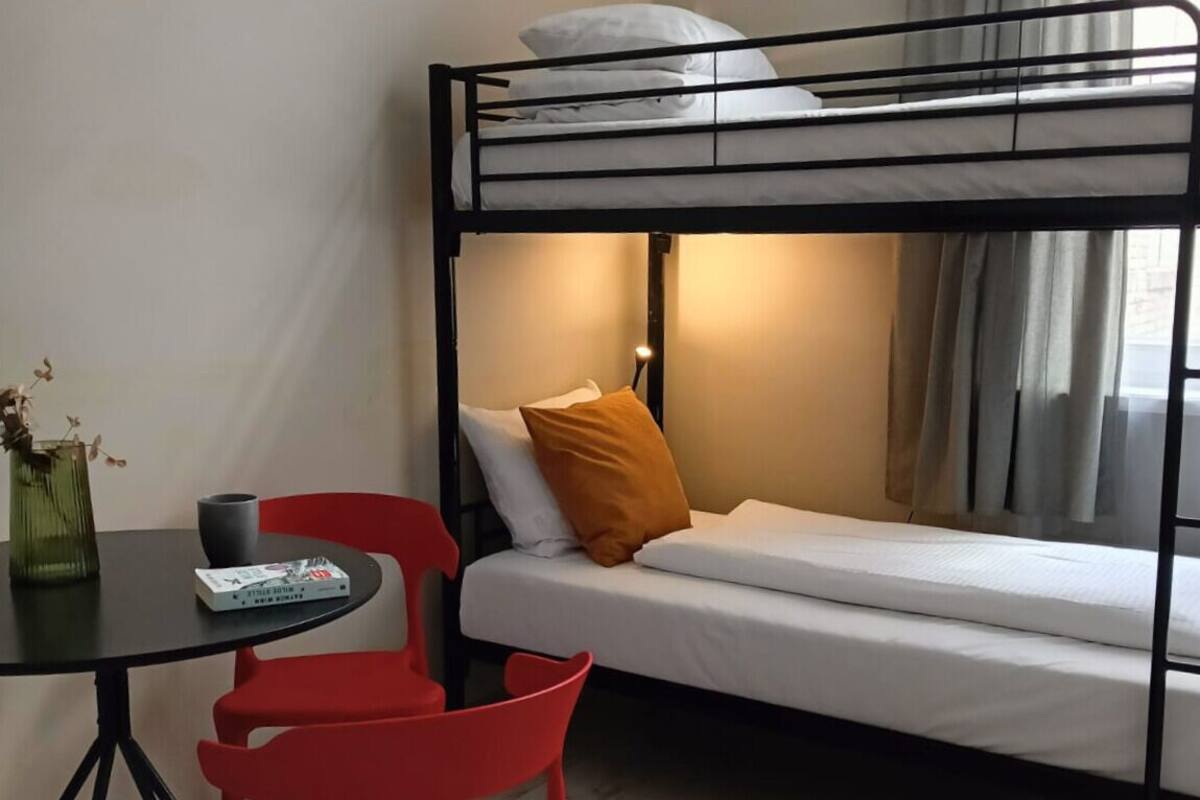 A bunk bed with a white mattress and a single orange pillow. There is a small table with a red chair and a vase with dried flowers.