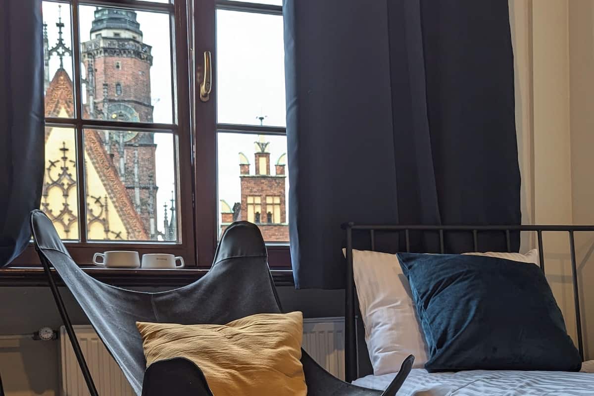 A room with a view of a historic building through a window, a black metal bed frame, a black folding chair, and a small rug on the floor.